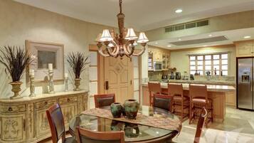 A dining area with a glass table, chairs, a chandelier, and a kitchen in the background.