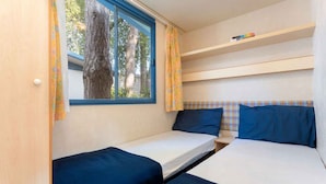 Quarto pequeno com duas camas, janela com cortinas e uma estante com livros.