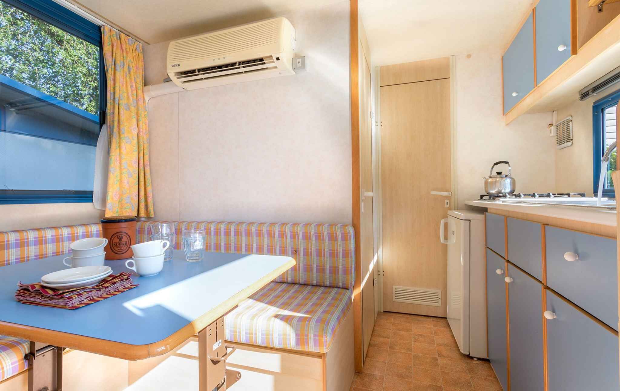 A compact kitchenette with a stove, sink, and a small refrigerator. There's a bench with a striped cushion and a table with a blue top.