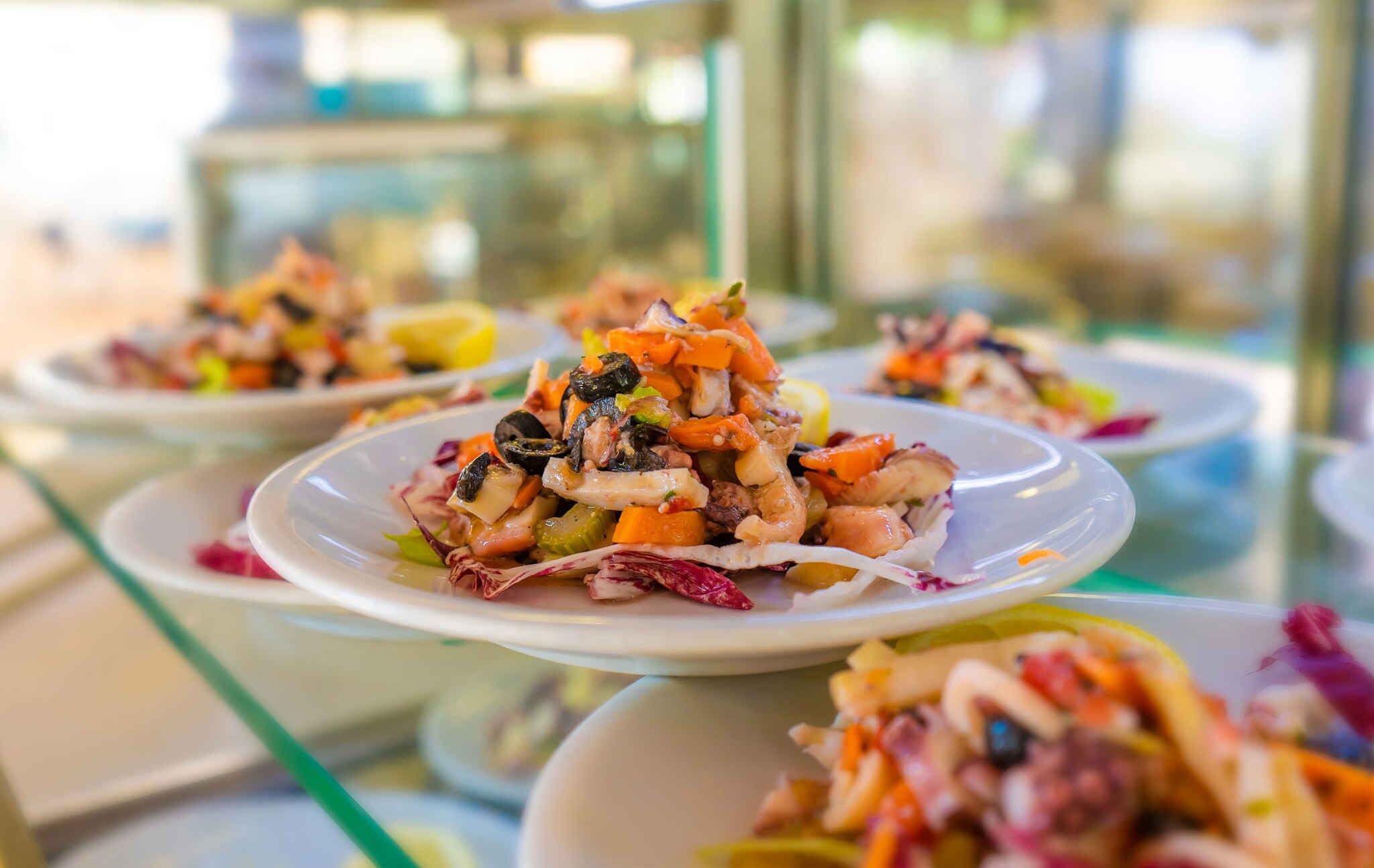 Buffet variado com diversos pratos, incluindo uma salada de frutos do mar com camarões, legumes e possivelmente outros frutos do mar.