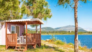 Una cabaña de madera con un porche, una mesa y sillas, con vistas a un lago y montañas al fondo.