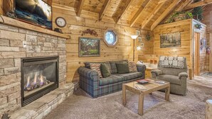 A cozy log cabin interior with a stone fireplace, a plaid sofa, a wooden coffee table, and a small armchair. - Mountain Eden, Hot Tub, Resort Pool, WIFI, $560 FREE tickets each paid day!!! (Gatlinburg)