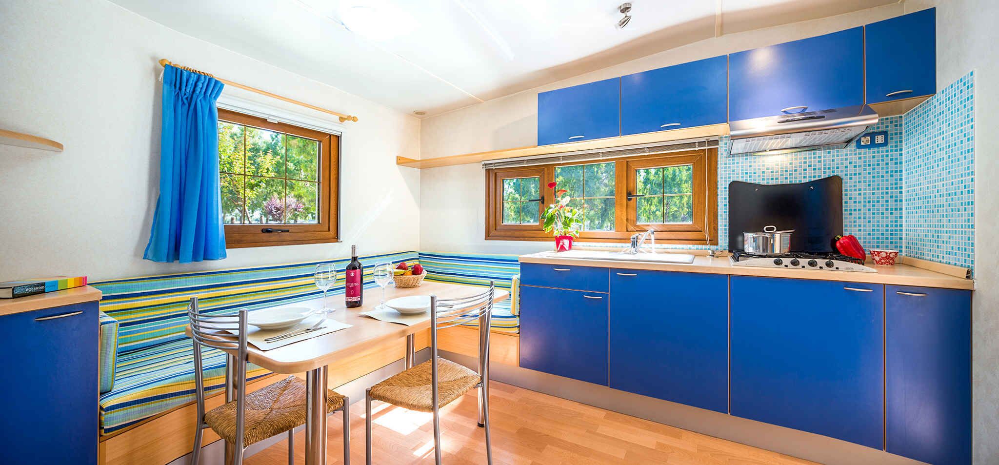 A modern kitchen with blue cabinets, a wooden dining table, and a window with a view of greenery.