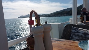 Taula amb saleires i pebreres, una ampolla i una cistella teixida sobre el pont d'un vaixell, amb vista al mar i a les muntanyes llunyanes.