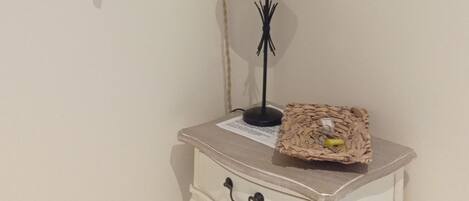 A small room with a white wall, a framed picture, a lamp, a table with a basket, and a wall-mounted light fixture.