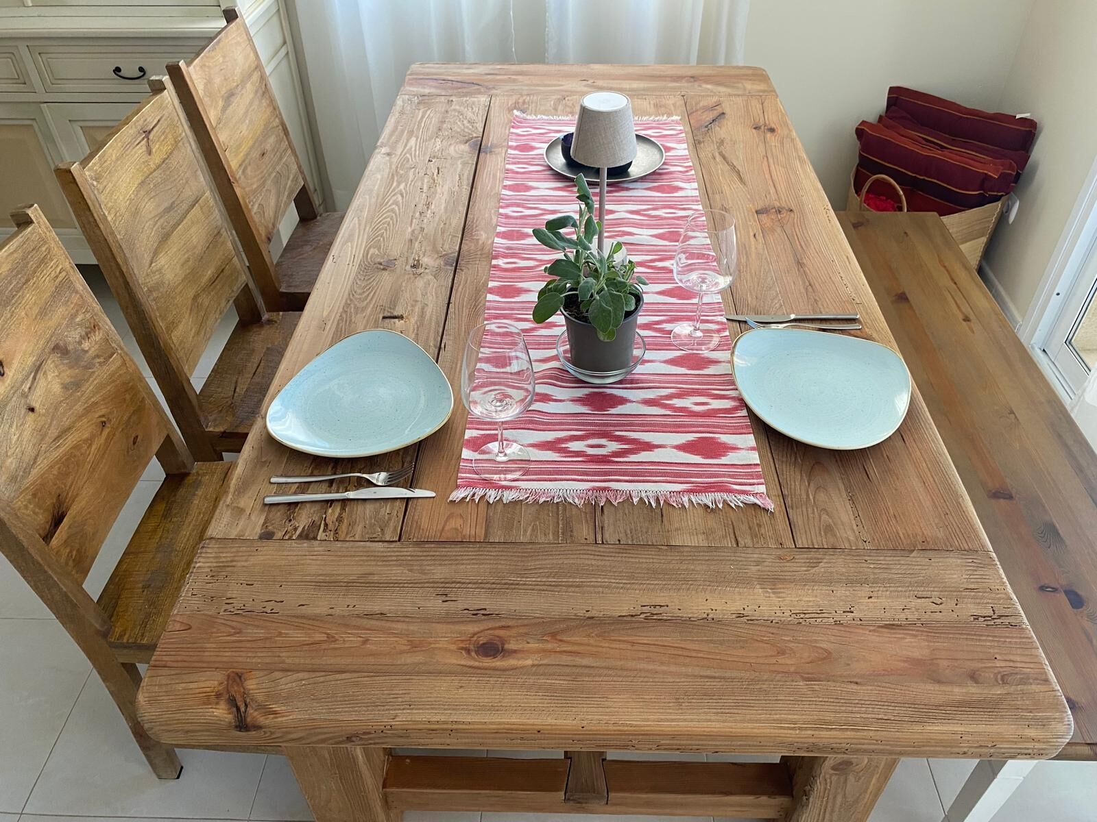 A wooden dining table with two plates, a glass, a potted plant, and a patterned placemat.