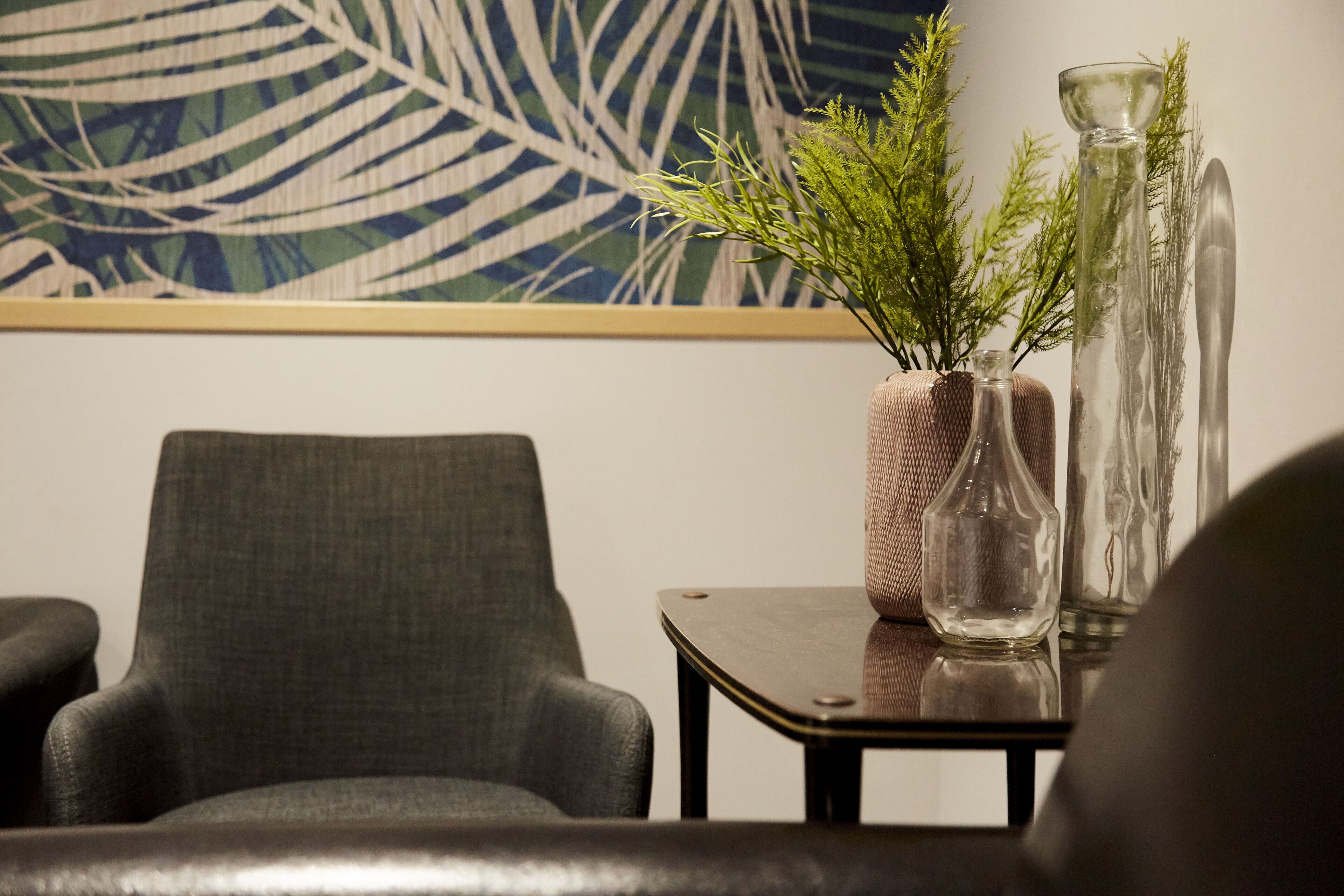 Une pièce comprenant un fauteuil à revêtement gris, une petite table sur laquelle est posé un vase rempli de feuillage vert, et un grand vase en verre.