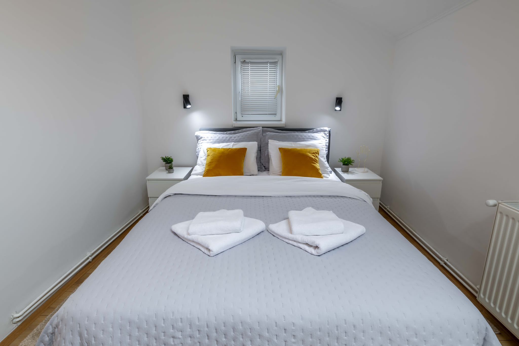 Una cama bien hecha con ropa blanca y dos almohadas, dos toallas blancas dobladas sobre la cama, una mesita de noche con una planta y una ventana con persianas.