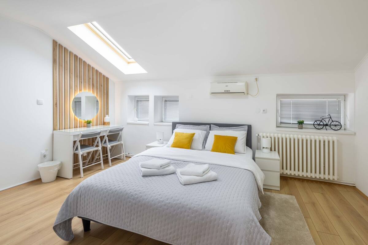 A modern bedroom with a skylight, a desk, a bed with white and yellow bedding, and a radiator.