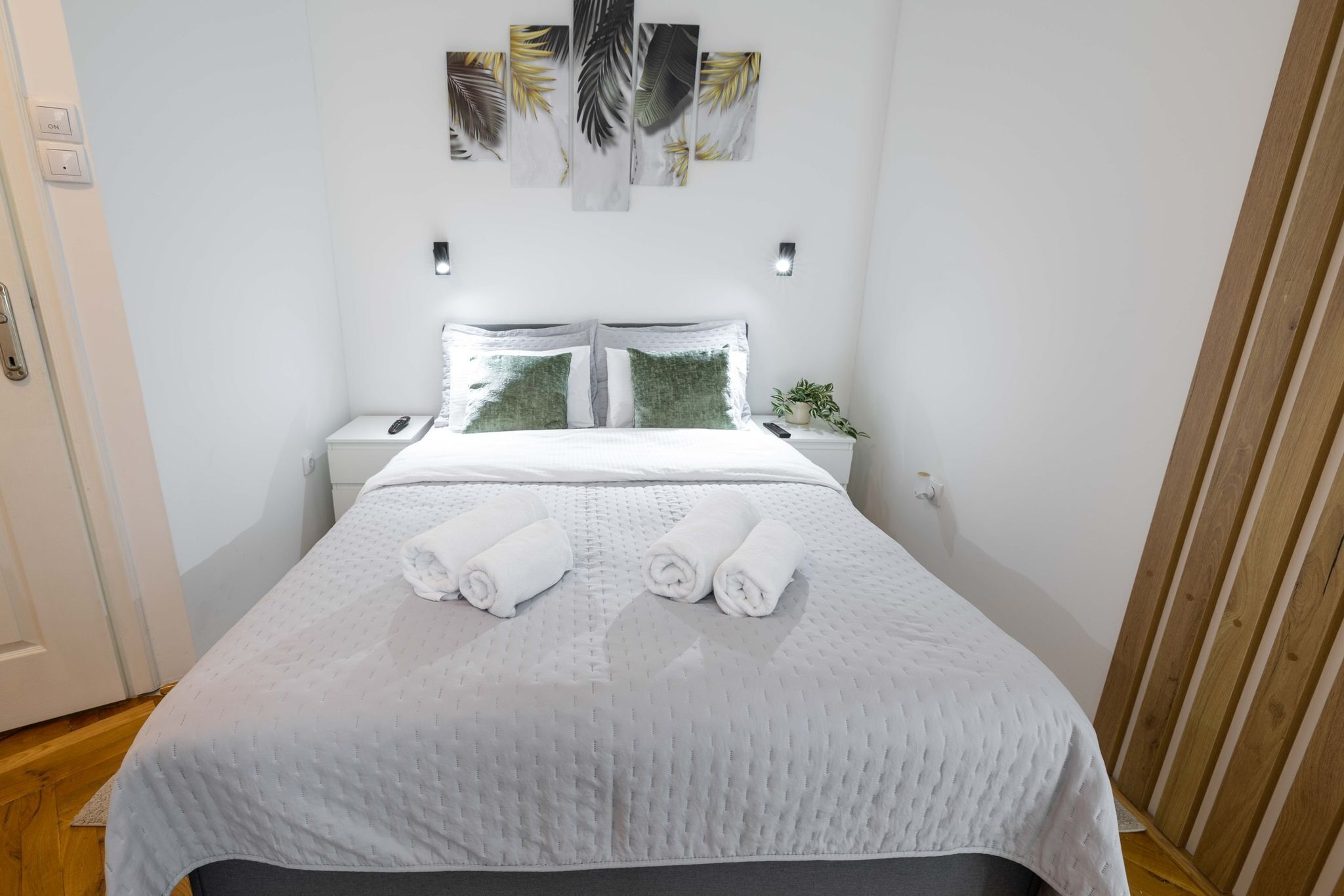 A neatly made bed with white linens and two rolled towels, a bedside table with a plant, and a wall with a leaf-patterned artwork.