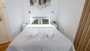 A neatly made bed with white linens and two rolled towels, a bedside table with a plant, and a wall with a leaf-patterned artwork.