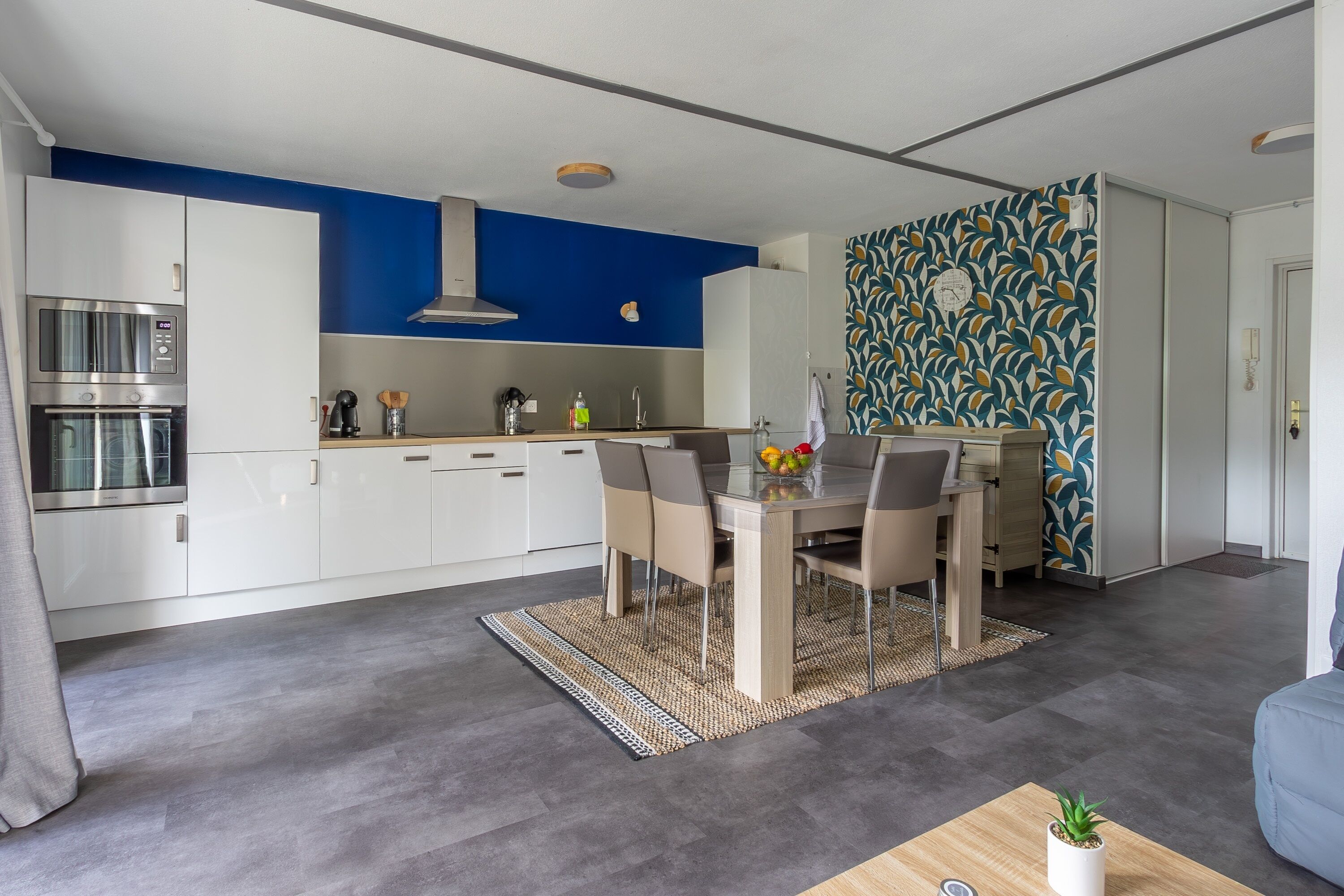 Een moderne keuken met witte kastjes, een blauwe accentmuur en een eethoek met een tafel en stoelen.