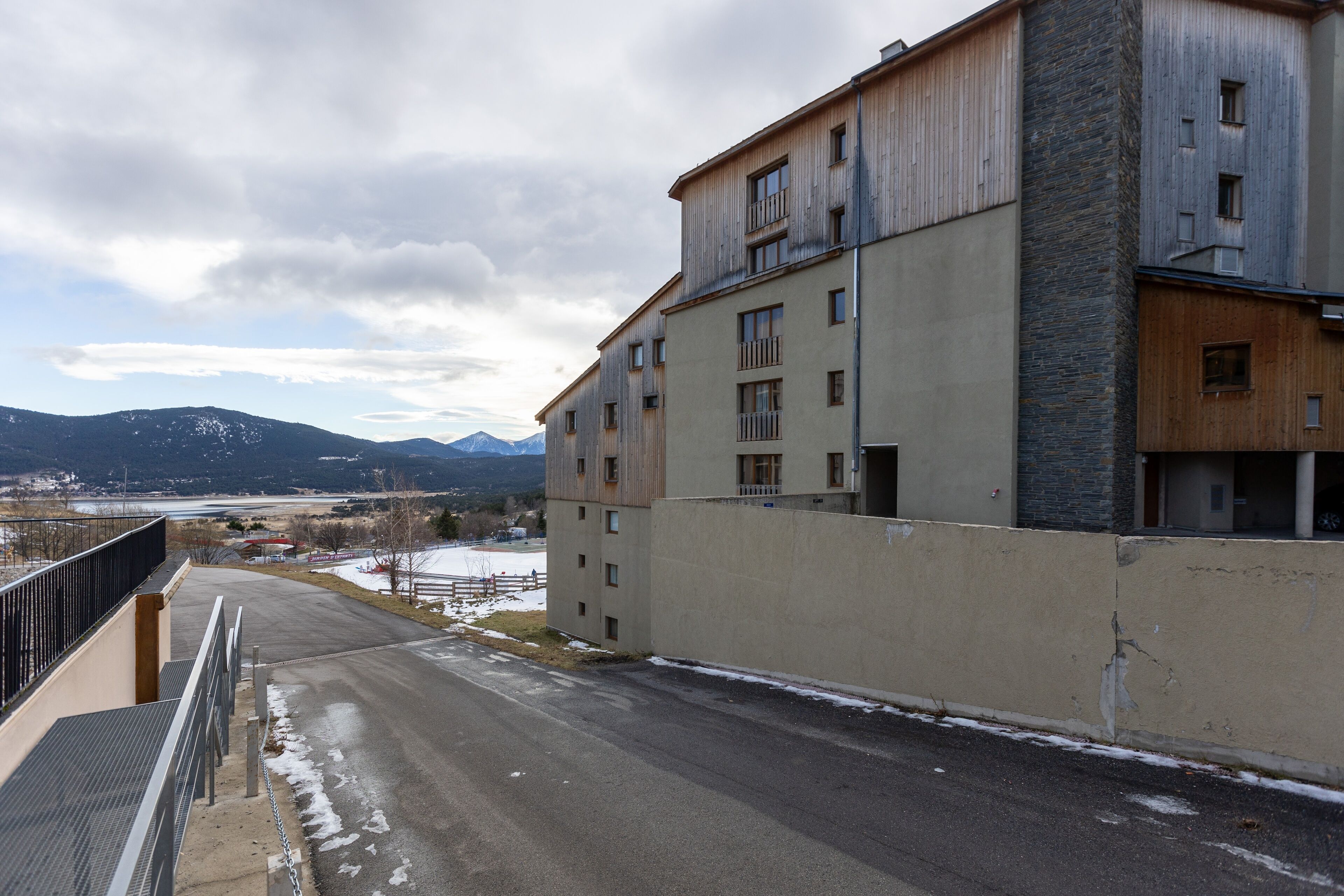 A multi-story building with balconies, a paved road in front, and mountains in the background.