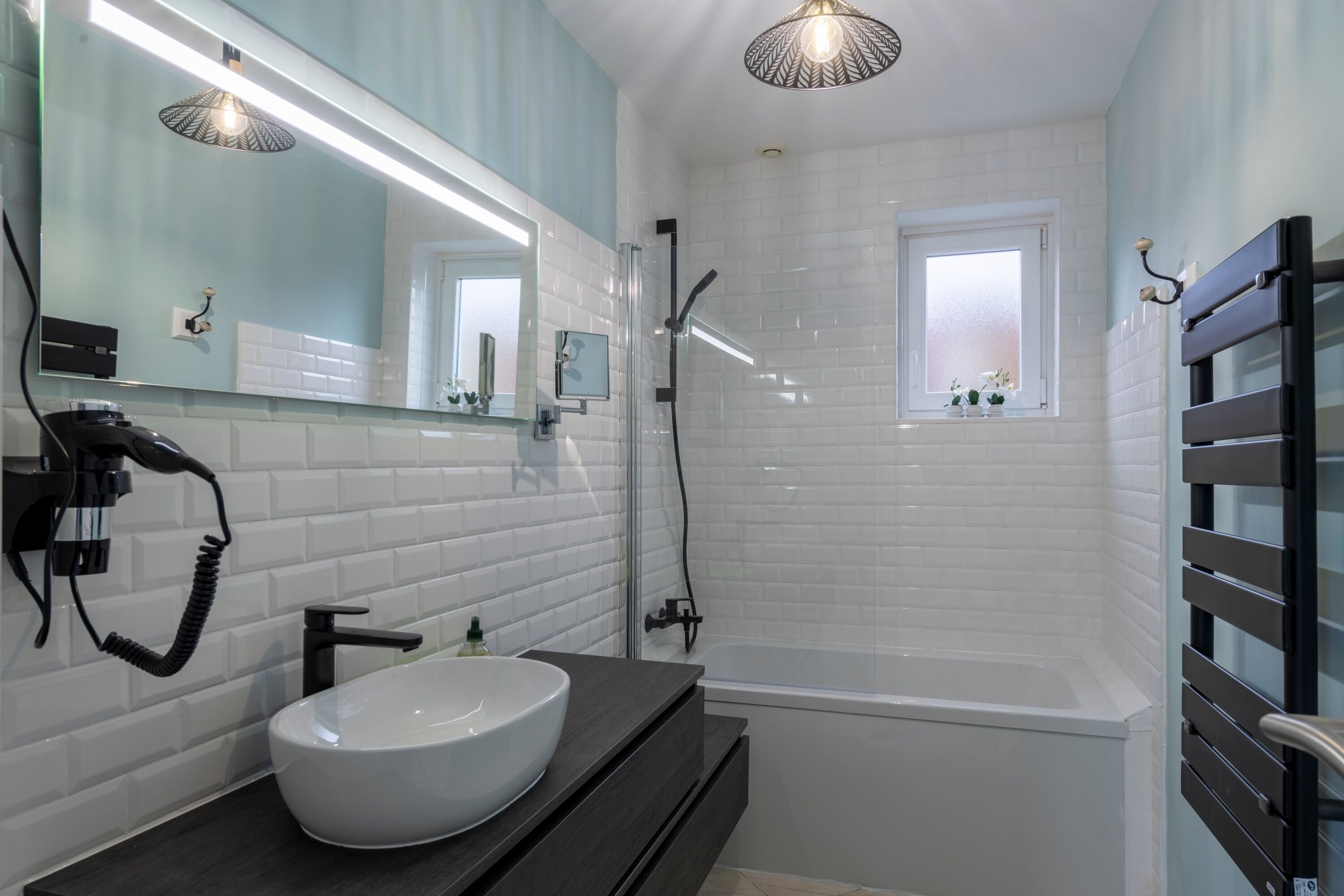 Un baño moderno con pared de azulejos blancos, un lavamanos grande en blanco, un mueble de tocador negro, una ducha con mampara de vidrio y una ventana con vistas al exterior.