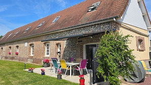 Ein Backsteinhaus mit geneigtem Dach, einer überdachten Terrasse mit Gartenmöbeln und einer kleinen Gartenanlage mit Topfpflanzen.