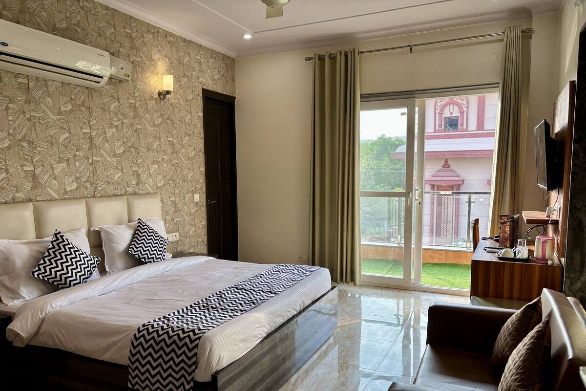 A hotel room with a large bed, a sofa, a TV, and a balcony with a view of a building.
