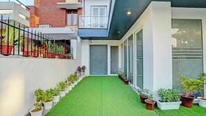 Um edifício moderno com telhado plano, janelas amplas e um caminho de grama sintética ladeado por plantas em vasos.