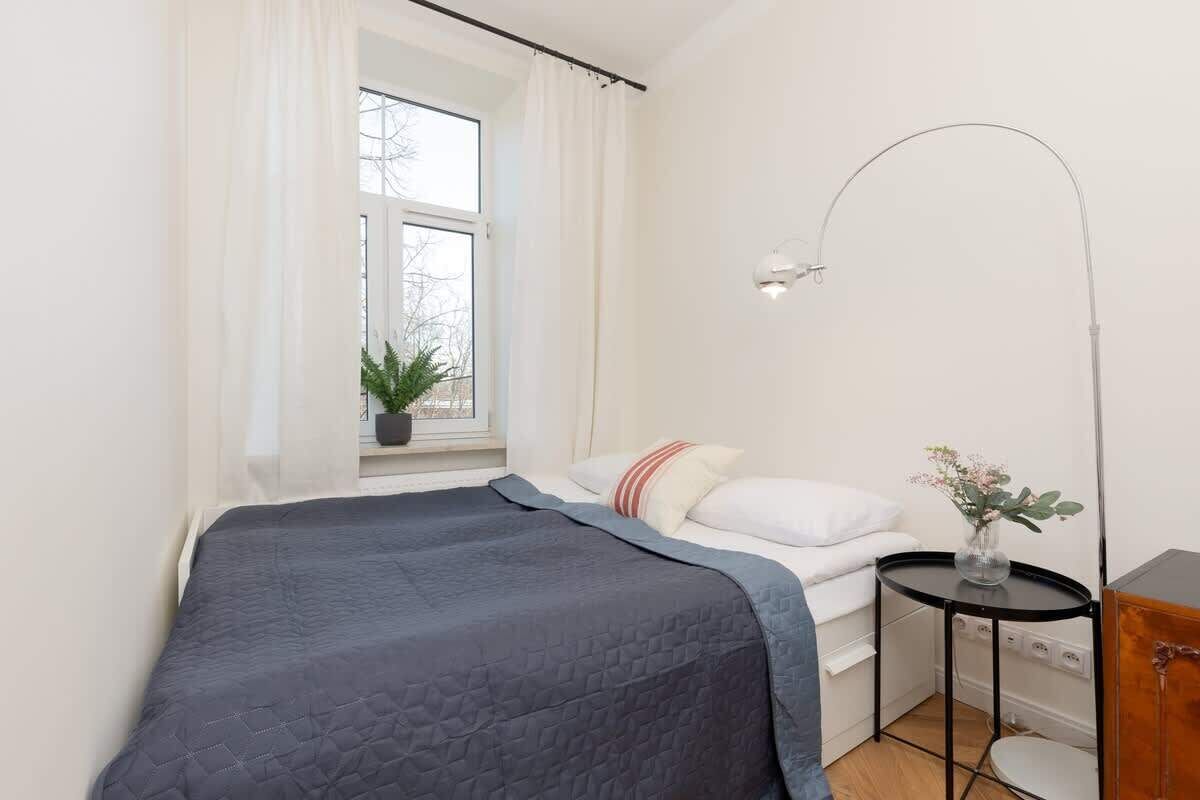A bedroom with a bed, a bedside table with a lamp, a window with curtains, and a potted plant.