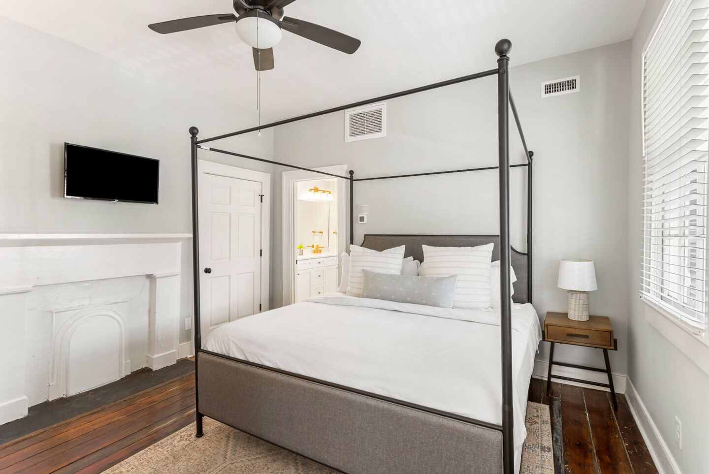 A four-poster bed with a headboard, a television mounted on the wall, a fireplace, and a window with blinds.