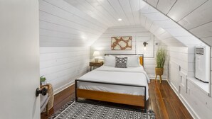 A bedroom with a sloped ceiling, a bed with white bedding, a wall-mounted lamp, a decorative wall piece, and a potted plant.