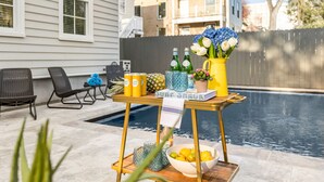 A poolside setup with a table holding drinks, a bowl of fruit, and a vase of flowers.
