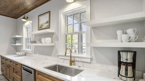 A kitchen with a white countertop, stainless steel sink, and a coffee maker. There are wooden cabinets and a window with a view of the outside.