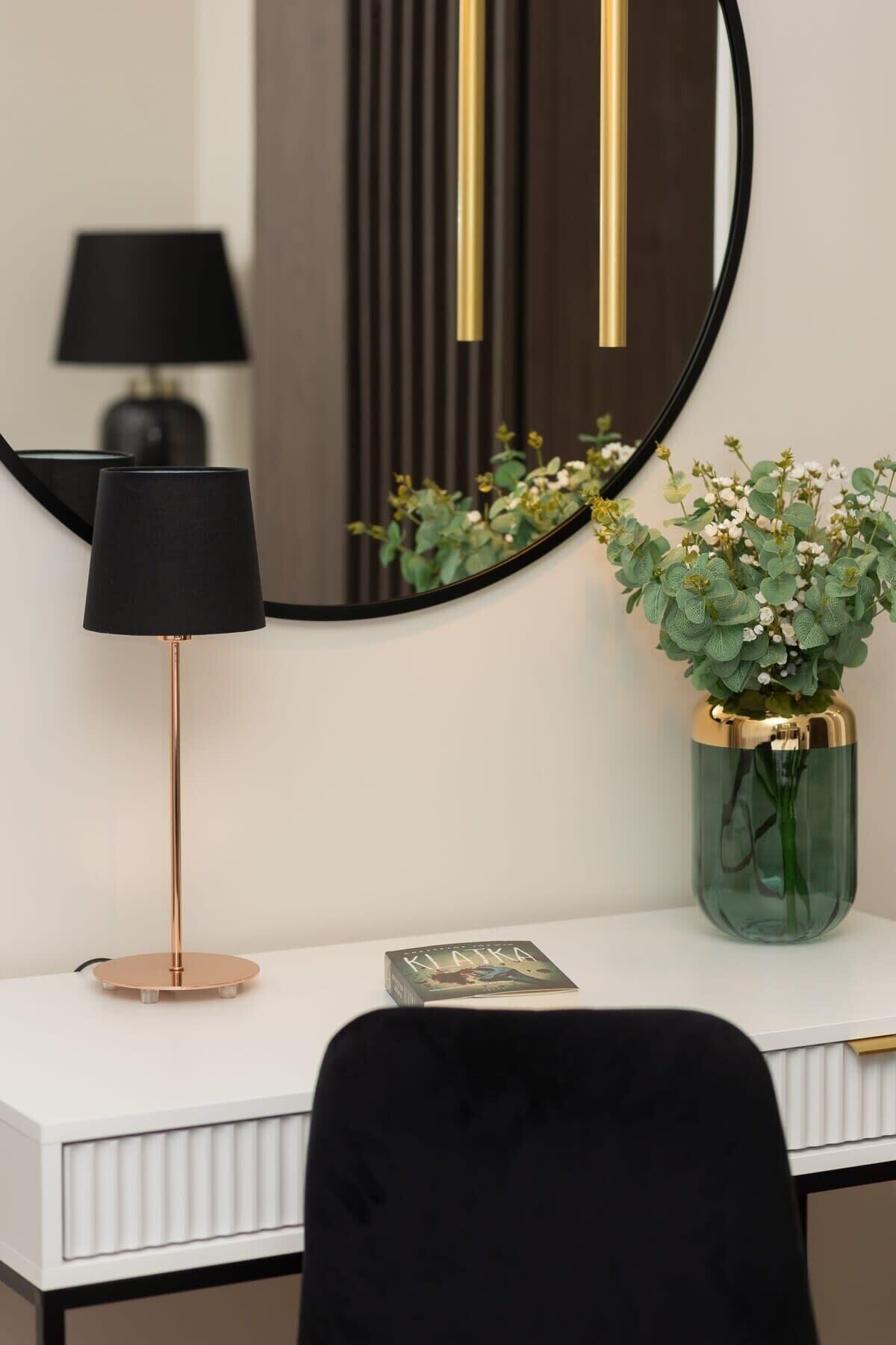 A modern desk with a black lamp, a round mirror, and a vase with green foliage.
