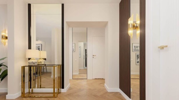 A hallway with a glass and gold console table, a mirror, and a door leading to another room.