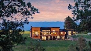 A modern two-story house with a large deck and multiple windows, surrounded by a grassy landscape and trees.