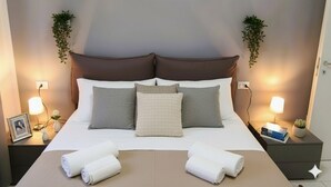 Una cama bien hecha con ropa de cama blanca y gris, dos mesitas de noche con lámparas, y una foto enmarcada sobre la mesa de la izquierda.
