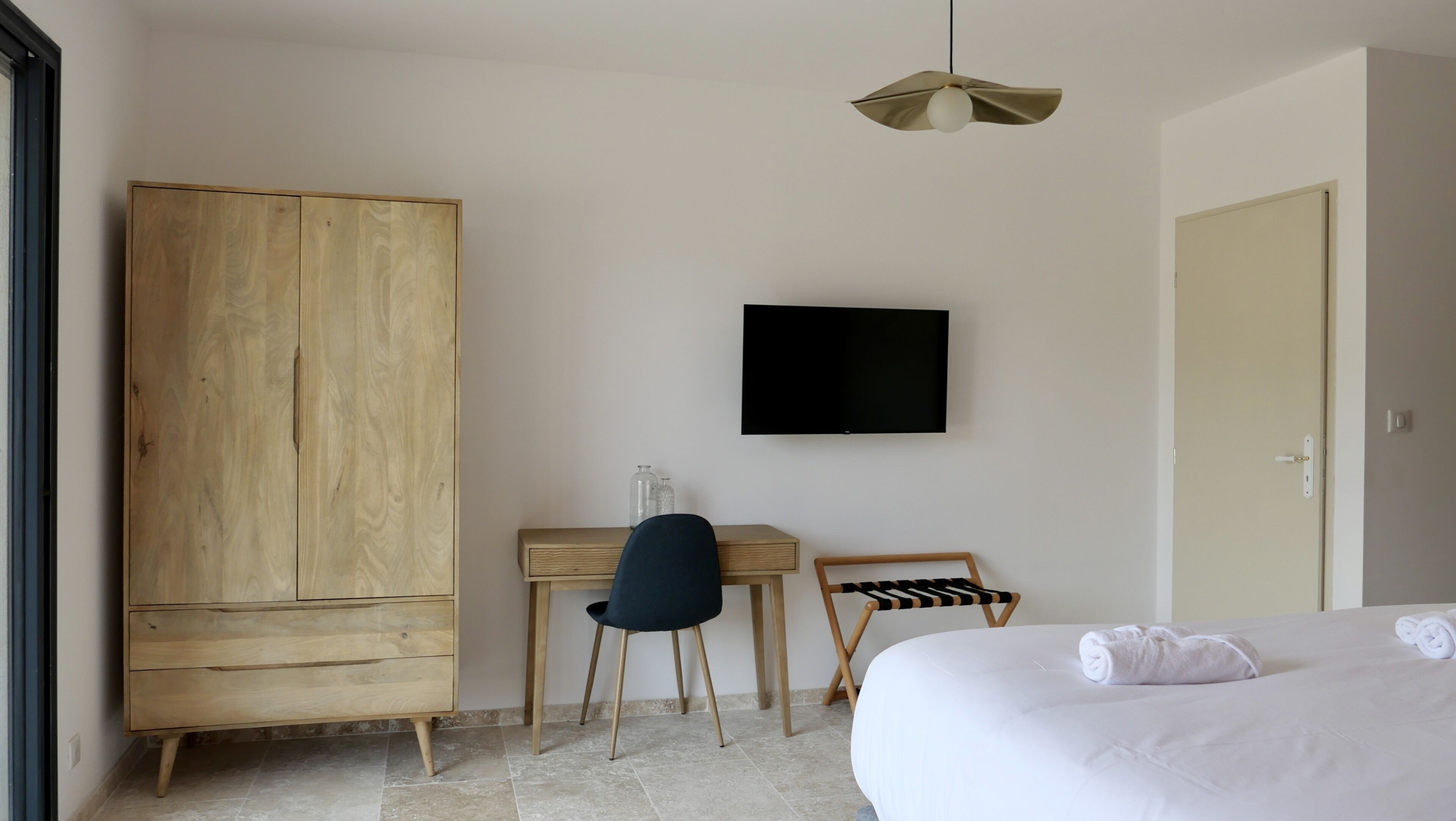 A hotel room with a wooden wardrobe, a desk with a chair, a television mounted on the wall, and a bed with towels.