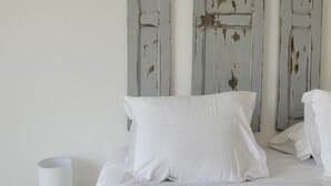 Una cama con sábanas blancas, una almohada y una toalla doblada sobre una mesita de noche de madera con una lámpara blanca.