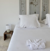 Una cama con sábanas blancas, una almohada y una toalla doblada sobre una mesita de noche de madera con una lámpara blanca.