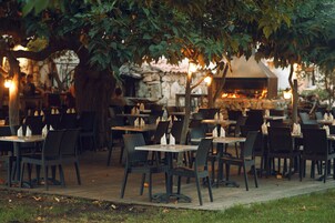 Un área de comedor al aire libre con mesas y sillas, un muro de piedra y un horno de leña al fondo.