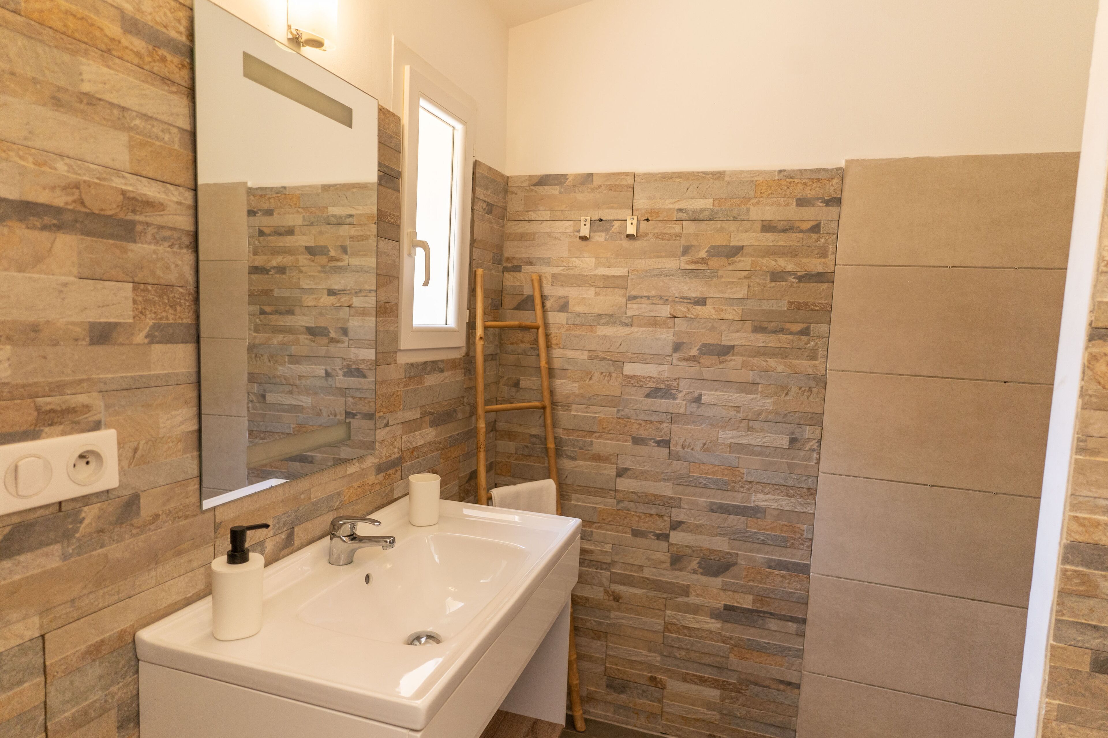 A bathroom with a stone wall, a white sink, a mirror, and a wooden ladder.