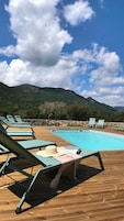 Una terraza de madera con sillones de descanso y una piscina, rodeada de montañas y un muro de piedra.