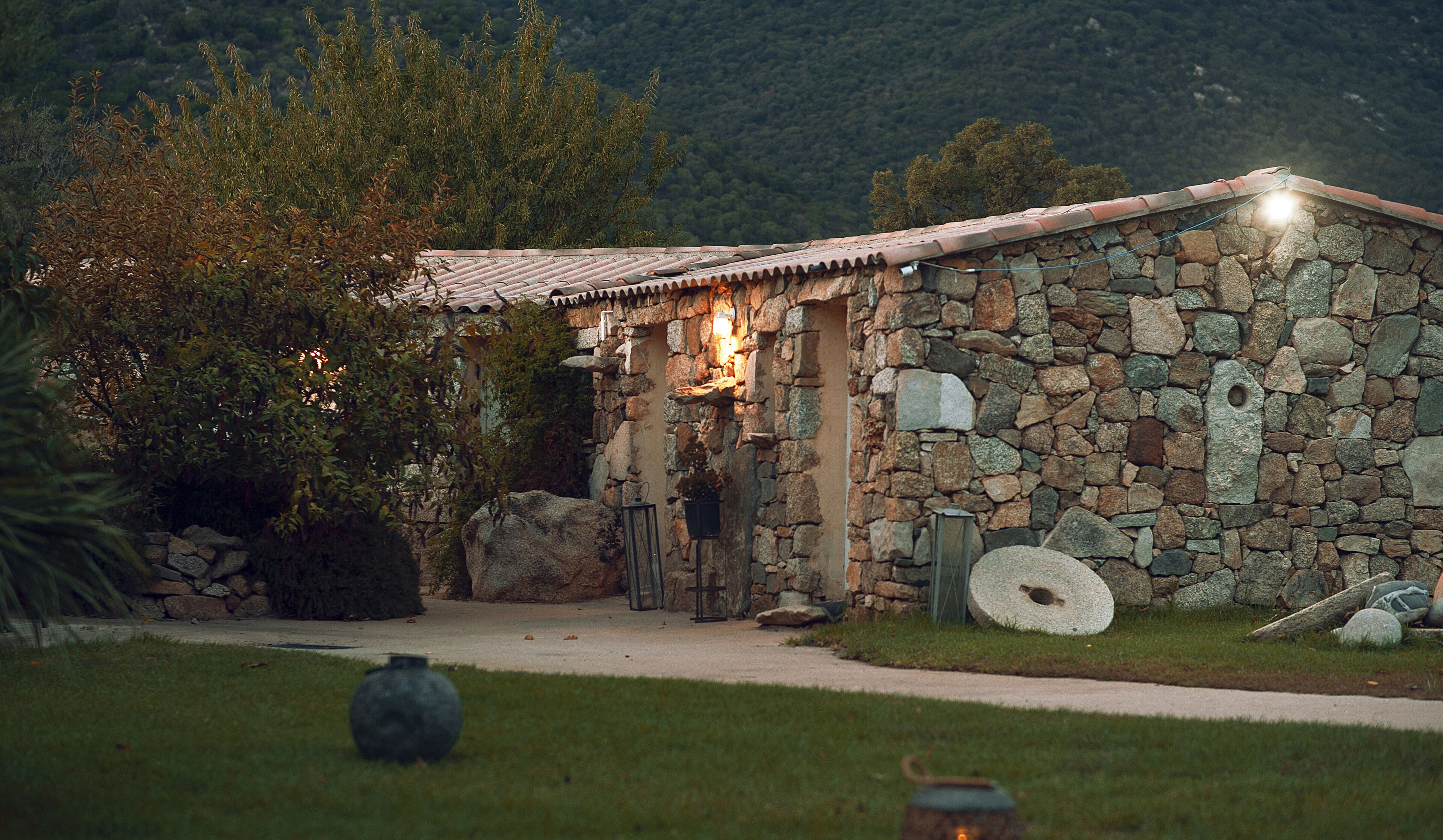 Un edificio in pietra con tetto in tegole, una parete in pietra e un piccolo giardino con una pianta in vaso e un lampione.