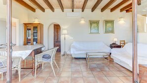 A room with a dining table, wicker chairs, a sofa, and a bed. There are two wall-mounted lamps and a painting on the wall. - S'Albufereta by Rentallorca (Alcúdia)