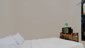 A single bed with white bedding, a small wooden nightstand with a television and a fan, and a window with a curtain. - OYO Life 92709 Kost Teras Cikapundung Syariah (Bandung)