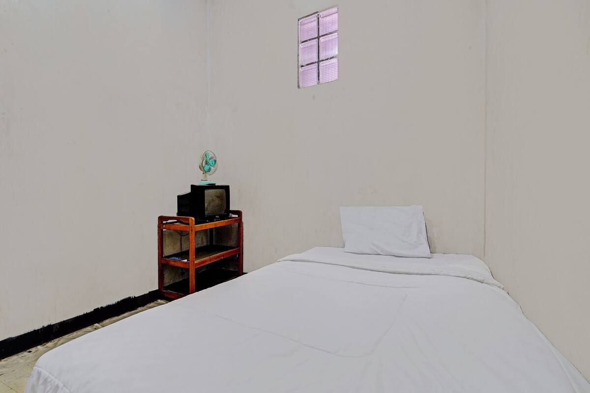 A room with a bed, a wooden nightstand with a television, a window, and a wall-mounted fan.