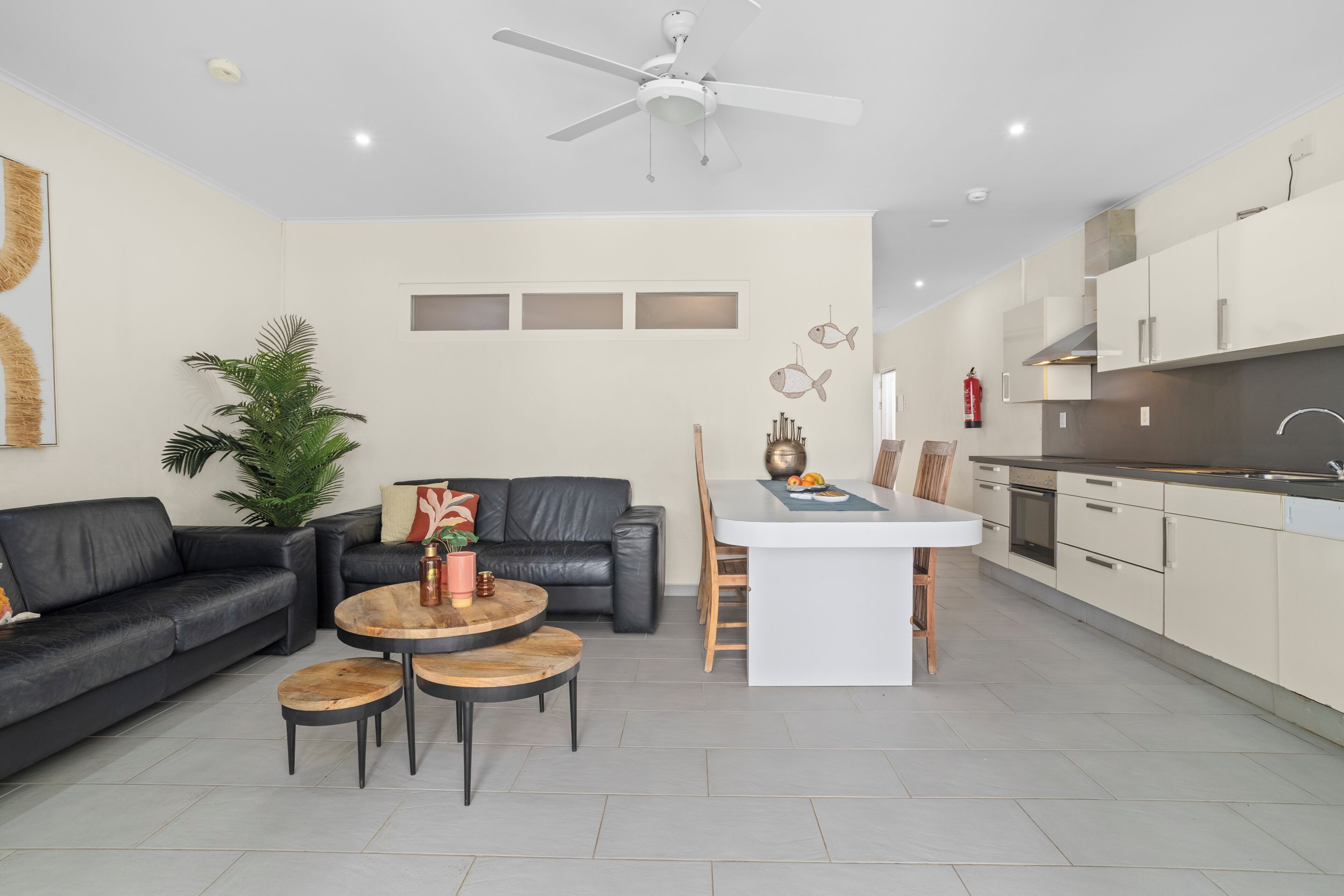 A modern kitchen with white cabinets, a central island, and a dining area with a black leather couch and wooden tables.
