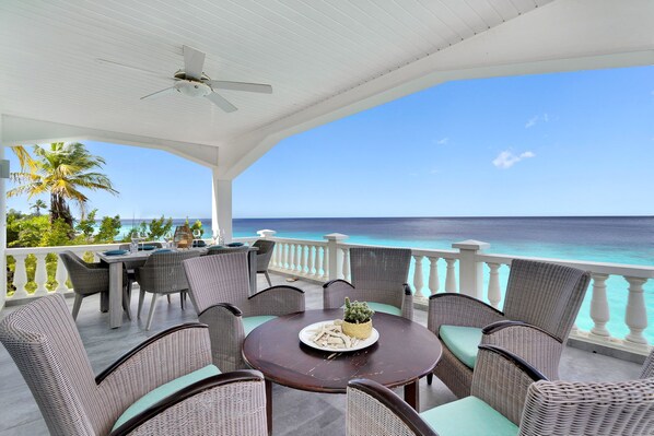 A covered outdoor seating area with wicker furniture and a table set for dining, overlooking a turquoise sea.
