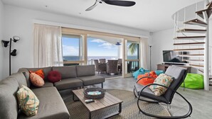 Ein modernes Wohnzimmer mit einer Couch, einem Couchtisch und einem Fernseher. Ein Balkon bietet Blick auf den Ozean.