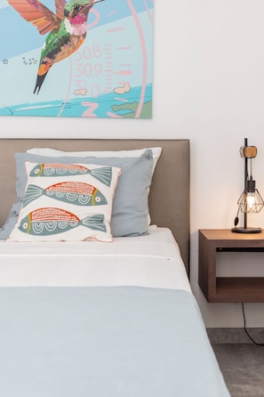 A bed with two pillows, one with a fish pattern and another plain, a nightstand with a lamp, and a wall with a colorful painting of a hummingbird.