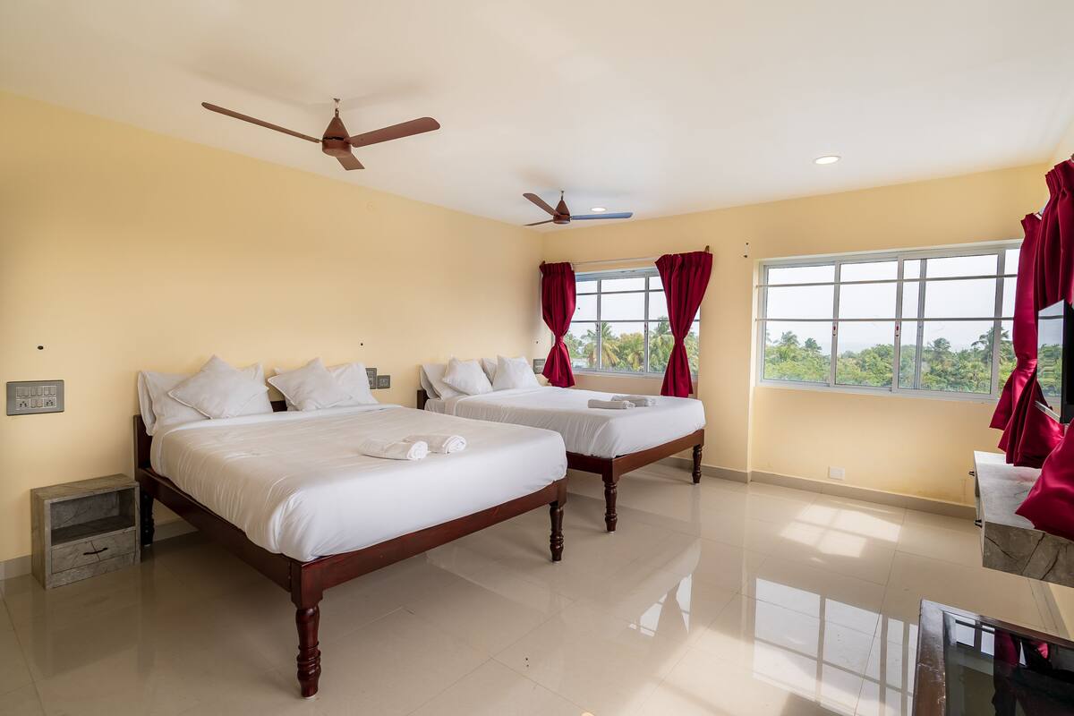 Habitación de hotel con dos camas, ventilador de techo, ventana con cortinas y vistas a la vegetación.