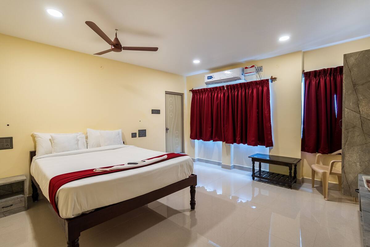 Una habitación de hotel con cama, ventilador de techo, equipo de aire acondicionado y ventana con cortinas rojas.