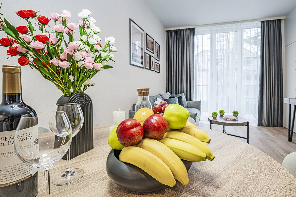 Een eetgedeelte met een tafel gedekt met fruit, wijn en glazen. Er staat een vaas met bloemen, een bank en een televisie op de achtergrond.