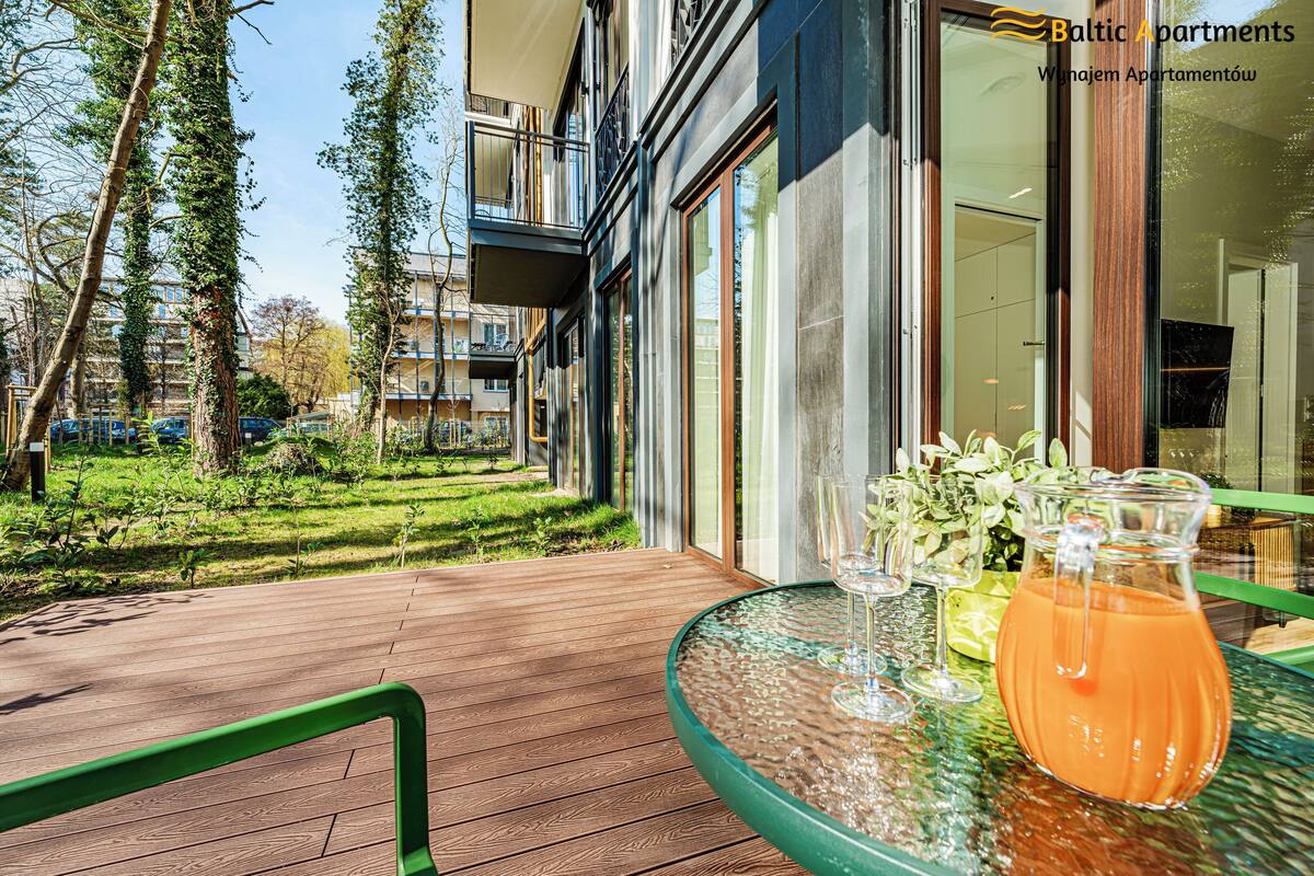 Een glazen tafel met een karaf sinaasappelsap, een glas water en een glas wijn op een balkon met een houten vloer en een groene balustrade.
