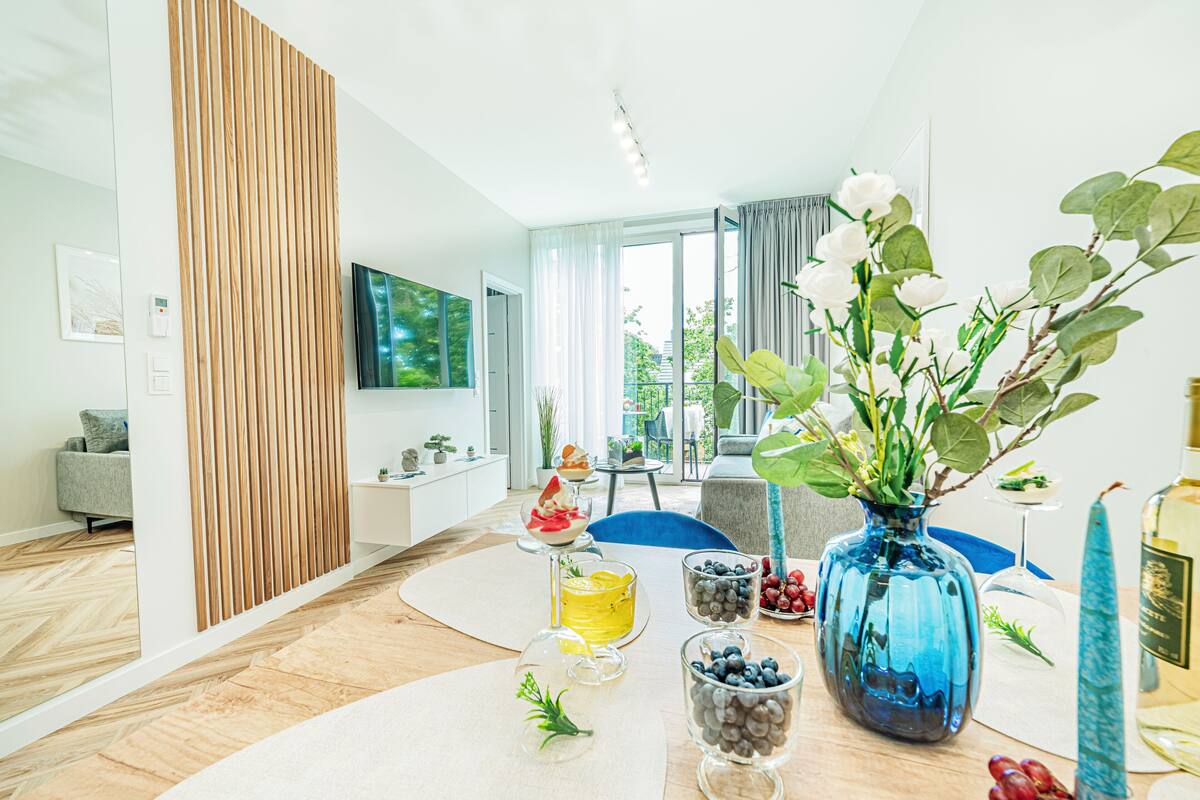 Een moderne woonkamer met een houten scheidingswand, een eettafel met fruit, een blauwe vaas en een aan de muur bevestigde televisie.