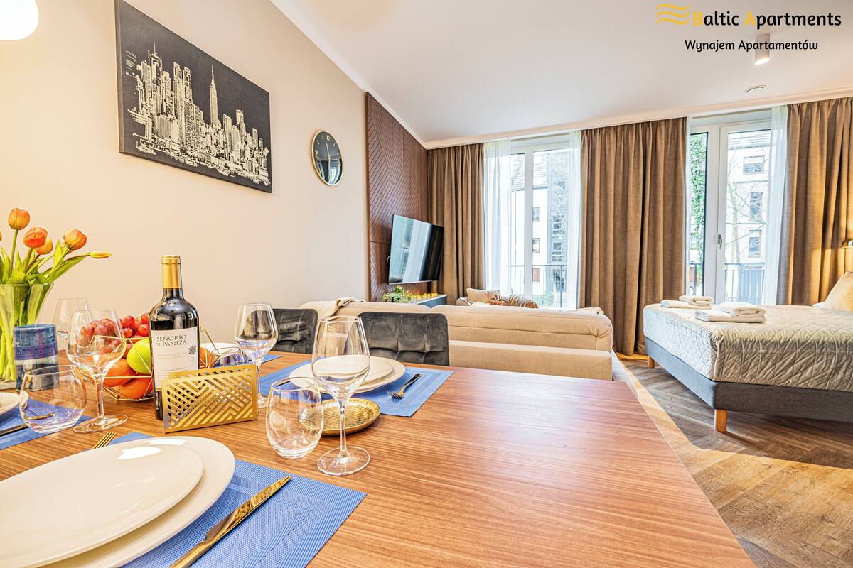Een goed verlichte hotelkamer met een eethoek gedekt voor een maaltijd, een bed, een televisie en grote ramen met gordijnen.